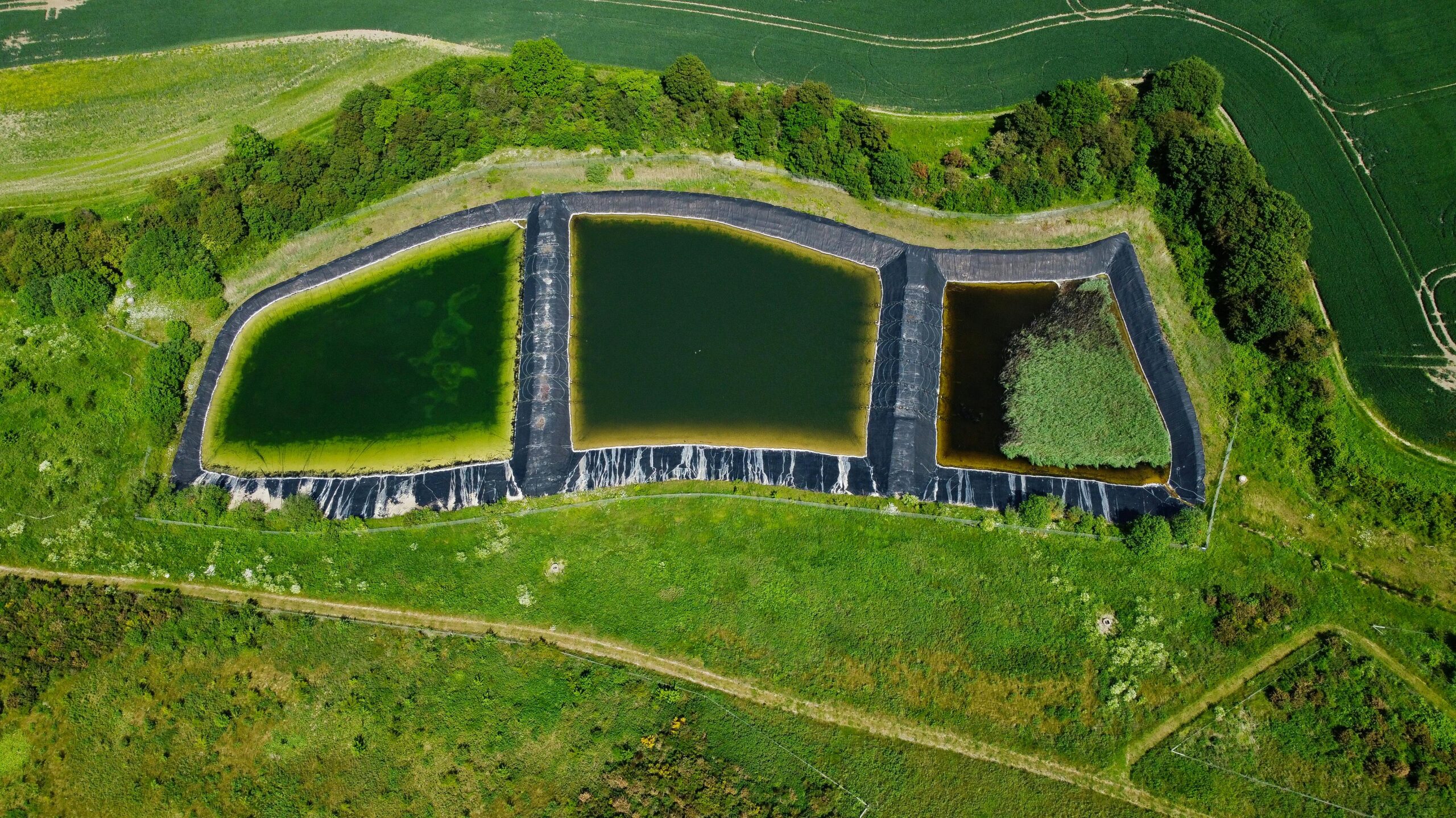 Aerial drone view of three black-lined, interconnected industrial ponds containing different stages of water treatment or storage, surrounded by lush green fields and trees.