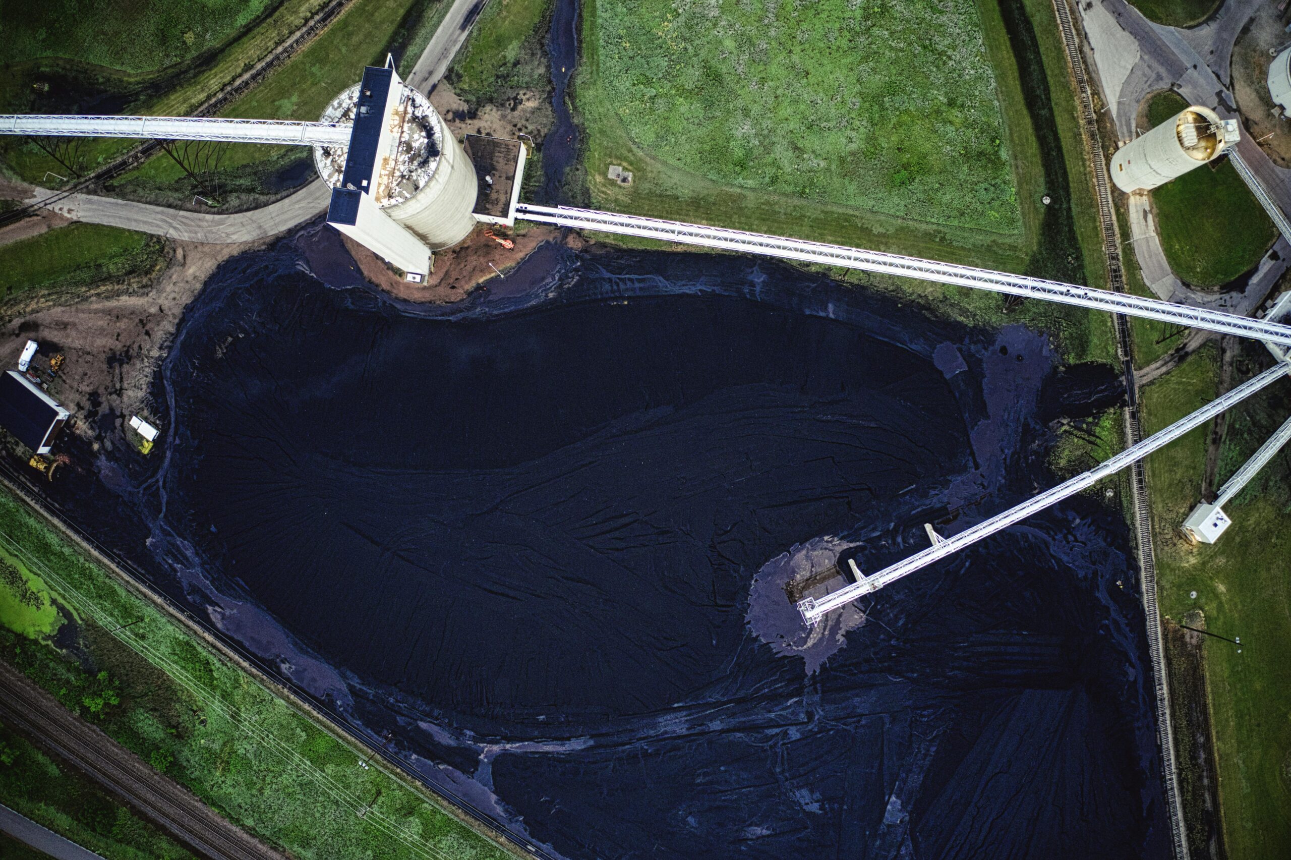 High-angle aerial view of a massive black coal or ore stockpile surrounded by conveyor belts, industrial silos, and green fields.